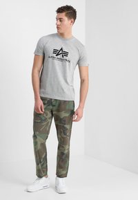 Grå bomulds t-shirt med sort "Alpha Industries" logo, parret med grønne stofbukser med camouflagemønster og hvide sneakers. Afslappet opsætning.