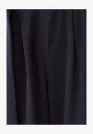 Primo piano di tessuto scuro con pieghe sottili e texture, che mostra materiale drappeggiato in un ambiente neutro.