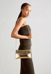 Metallic gold clutch bag with a geometric design and flat shape, featuring a chain strap. Worn with a ribbed dark brown dress.