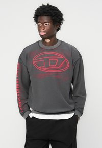 Sweatshirt i mørkegrått stoff med rund hals. Har rødt logo og tekst på brystet og ermet. Brukt over en hvit skjorte.