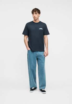 Young man wearing a navy blue graphic T-shirt, loose light blue jeans, and black lace-up sneakers, standing with one hand in pocket.