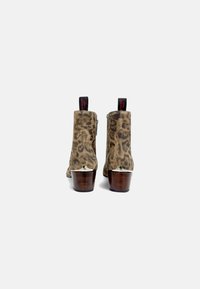 Botas de ante estampadas de leopardo hasta el tobillo con un tacón de bloque de madera, cremallera trasera y una etiqueta para tirar en rojo. Los acentos incluyen un detalle plateado en el tacón.