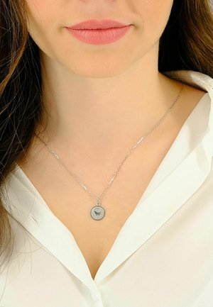 Collana con catena d'argento e pendente circolare con un disegno di bisonte, indossata sopra una camicia bianca. Il pendente ha una superficie liscia e lucida.