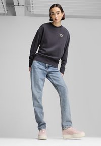 Grijze sweatshirt met lange mouwen en logo, lichtblauwe relaxte jeans en lichtroze sneakers met textuurzolen, tegen een neutrale achtergrond.