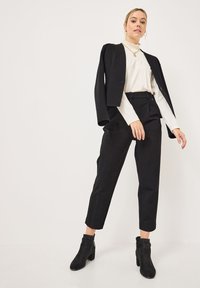 Blazer noir ajusté sur un haut blanc à col montant, associé à un pantalon noir cropped et des bottines, avec un design lisse et texturé.