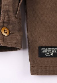 Smeđa pamučna tkanina s teksturiranim završetkom, s velikim zlatnim gumbom i crnom etiketom s bijelim tekstom za Rivvia Projects.