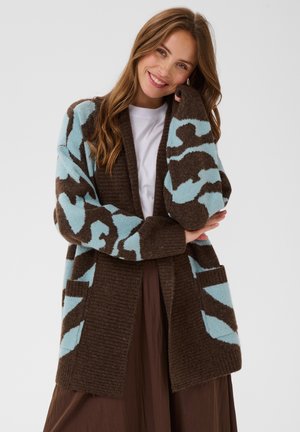 Femme souriante aux longs cheveux portant un cardigan marron à motif abstrait bleu clair, une chemise blanche et une jupe marron, debout devant un fond uni.