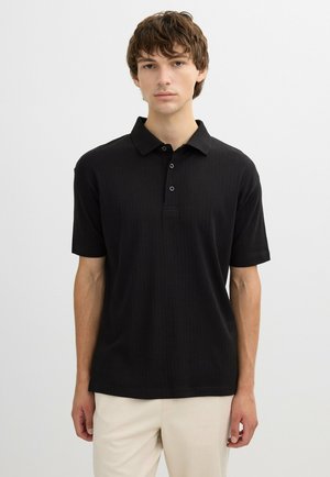 Joven con una camisa polo negra de manga corta y pantalones beige claros, de pie contra un fondo blanco liso.