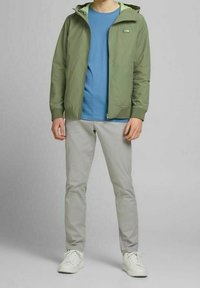 Giacca a vento verde con cappuccio sopra una t-shirt blu, abbinata a pantaloni grigi e sneakers bianche. La giacca presenta una chiusura con zip ed è leggera.