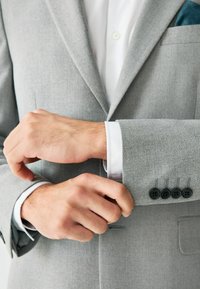 Blazer gris con tejido texturizado, que incluye una camisa blanca debajo. Los puños tienen cuatro botones negros y un pañuelo decorativo en el bolsillo.