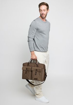 Man in gray sweater and cream pants walking while holding a brown briefcase-style bag with front pockets and handles.