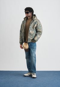 Pufnasta jakna s kapuljačom u kamuflažnom uzorku, smeđa majica, plave traperice, bež kapa i siva obuća za trčanje s crnim vezicama, stoji na plavom tepihu.