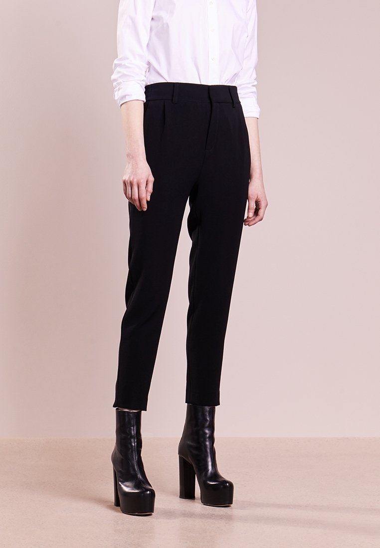 Femme portant un pantalon noir ajusté coupé à la cheville, une chemise blanche à manches longues et des bottines plateformes noires, debout sur un sol beige.
