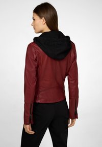 Veste en cuir rouge avec capuche noire. Les caractéristiques incluent des poignets zippés, une coupe ajustée et une texture lisse, présentée de dos.