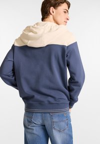 Sudadera color bloqueada en azul marino y crema con capucha ajustable, textura suave y dobladillo acanalado, combinada con jeans de mezclilla azul claro.