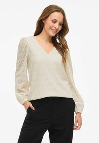 Kobieta uśmiechająca się, w beżowej bluzce z długim rękawem i dekoltem w serek oraz czarnych spodniach, z rękami w kieszeniach, na tle białym.