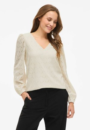 Femme souriante, portant un blouson beige à manches longues avec un col en V texturé et un pantalon noir, les mains dans les poches, sur fond blanc.