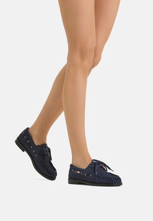 Gambe che indossano mocassini in camoscio blu navy con dettaglio stringato e suola nera, in piedi su uno sfondo bianco semplice.