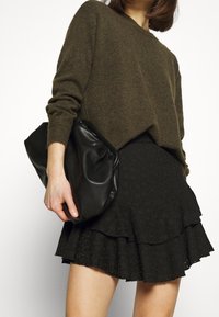 Pull en tricot vert olive avec un col rond. Jupe froncée noire avec des motifs floraux découpés. Pochette en cuir noir à la main.