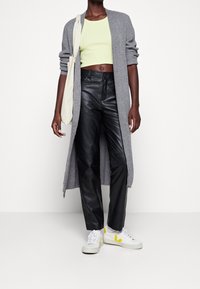 Long gilet en maille grise porté sur un top court vert clair, associé à un pantalon en cuir noir et des baskets blanches avec des accents jaunes.