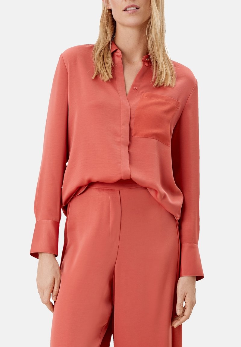 Blouse en soie corail avec une fermeture à boutons, un col classique et une poche poitrine unique. Coupe ample avec manches longues et accents de poignets.