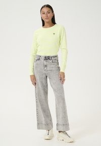 Langarm gestreepte shirt in neon geel en wit, met een klein groen hartembleem. Gecombineerd met high-waisted, lichtgrijze wijde jeans.