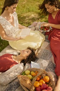 Trois femmes se détendent sur une couverture de pique-nique ; l'une lit, une autre tient un bol de fraises, et une est allongée les yeux fermés près d'un panier de fruits et des cartes.