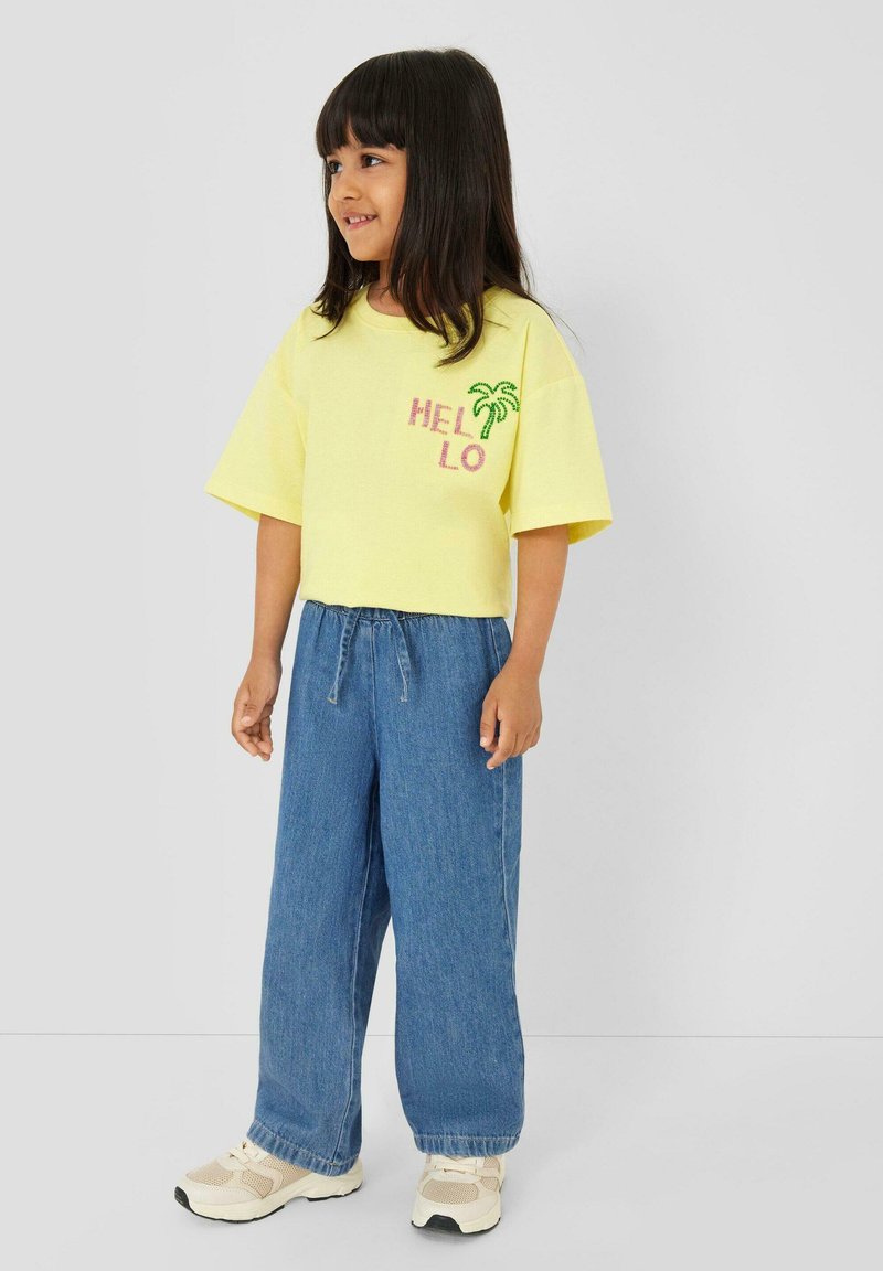 Niña joven con cabello negro, vistiendo una camiseta amarilla con la palabra "HELLO" y una palmera, jeans azules holgados y zapatillas cremas, de pie frente a un fondo liso.