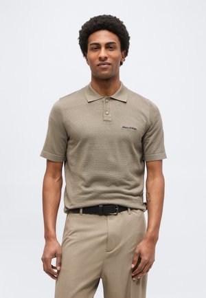 Άνδρας που φοράει μπεζ κοντομάνικο πόλο με το λογότυπο "Marc O'Polo", μπεζ παντελόνι και μαύρη ζώνη, στέκεται μπροστά σε ανοιχτό φόντο.