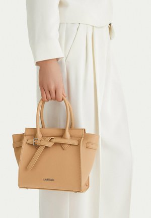 Mano sosteniendo un pequeño bolso de cuero beige con doble asa y detalle de cinturón, combinado con pantalones plisados blancos y blusa de manga larga.