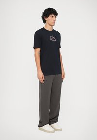 Camiseta de algodón negra con un logo "AX" en blanco y rojo en el lado izquierdo del pecho, combinada con pantalones anchos grises y zapatillas blancas.