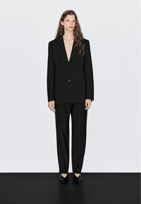 Zwart op maat gemaakt pak met een diepe V-hals blazer en brede broekspijpen. Soepele stof, minimalistisch ontwerp en puntige zwarte schoenen.