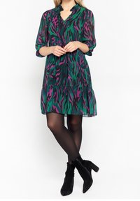 Šifonové šaty s leopardím vzorem v zelené a fialové barvě, s výstřihem do V, třičtvrtinovými rukávy a volánovým lemem. Nosené s černými kotníkovými botami.