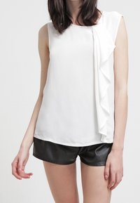 Weißes ärmelloses Oberteil mit einem Rüschendesign auf der linken Seite, kombiniert mit schwarzen Leder-Shorts. Glatte Textur, lässige Passform.