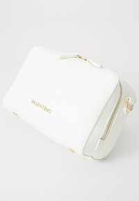 Vit texturerad läderväska med guldlogo, dragkedja och detaljer. Lätt böjd form med fyra guldfötter på basen. Kompakt design.