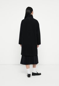 Zwarte wollen jas met hoge kraag, oversized vorm en verlaagde schouders, gecombineerd met een geplooide zwarte rok en zwarte schoenen met witte sokken.