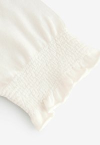 Bijela tkanina s smoktanim teksturom i detaljem volančića na rubu. Mekan, lagan materijal, elegantno skupljen na manžetama.