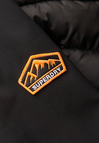 Sort stof med en struktureret overflade præsenterer et rektangulært orange og sort broderet patch med påskriften "SUPERDRY" og iskolde bjerggrafikker.