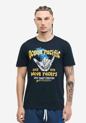 Hombre con cabello trenzado y tatuajes lleva una camiseta gráfica negra que dice "Ocean Pacific Wave Racers" con el gesto de mano "Good Vibes Only" y palmeras.