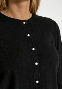 Svart cardigan i mjukt, texturerat tyg med rund halsringning och sex vita knappfästen framtill.
