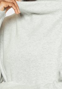 Suéter gris claro con una textura suave, que cuenta con un cuello alto y mangas largas, parcialmente sostenido por una mano.
