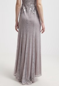 Femme portant une robe longue en dentelle violet clair avec une broderie florale sur le haut du dos, vue de dos.
