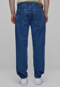 Modré džíny z denimu s uvolněným střihem, obohacené o dvě zadní kapsy a koženou nášivku na pase. Tkanina má mírně vybledlý vzhled.