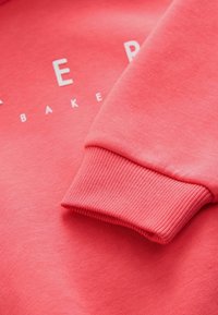 Felpa color corallo con polsini a coste e scritta bianca "BAKERY" sul davanti. Texture morbida e design semplice.