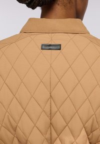 Giacca trapuntata beige con motivo a rombi, etichetta nera con "TRUSSARDI" e colletto. Texture liscia con vestibilità su misura.