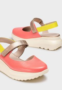 Zapatos de cuero color coral con punta al aire y suela gruesa color crema. Presentan una correa metálica y una correa trasera amarilla para mayor soporte.
