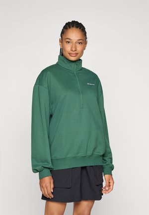 Grüner Pullover mit hohem Kragen, langen Ärmeln, Rippbündchen und Saum. Übereinander getragen über schwarzen Shorts. Logo auf der Brust. Weicher Stoff.