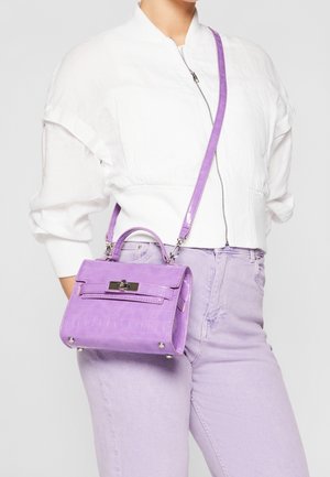 Kobieta ubrana w białą kurtkę na zamek i jasnofioletowe dżinsy, niosąca małą fioletową torebkę o fakturze skóry krokodyla z górnym uchwytem i paskiem na ramię.