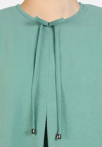 Turquoise blouse with a round neckline, featuring a keyhole opening at the front, tied with two thin strings ending in metal tips. Lightweight fabric.