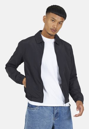 Mężczyzna w czarnej kurtce na zamek błyskawiczny noszonej na białej koszulce i niebieskich dżinsach, z ręką w kieszeni kurtki, stoi na tle jasnego, jednolitego tła.